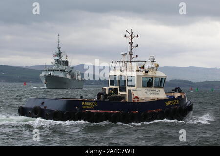 Bruiser, eine Damen Stan 1907 tug von Clyde Marine Services betrieben, Pässe Greenock mit hmcs St John's im Hintergrund. Stockfoto