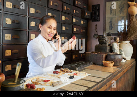 Lächelnd reife asiatische Frau suchen am grossen trockenen Berry in die Hand und am Telefon sprechen mit Client bei traditionellen Apotheke arbeiten Stockfoto