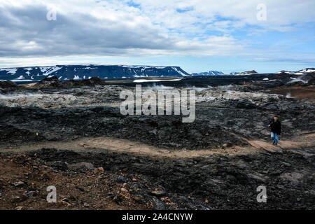 Leirhnjukur alten schwarzen Lavafeld mit einem unkenntlich Tourist auf Trail, schneebedeckten Bergen und blauem Himmel in Island, bewölkter Tag im Sommer, film Effekt Stockfoto