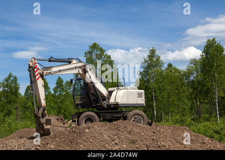 Ein weißer Mobilbagger steht auf einem Haufen von Schutt und Steine im Intervall zwischen den Arbeiten zur Instandsetzung einer Straße im Hintergrund einer Landschaft mit einem blauen Stockfoto
