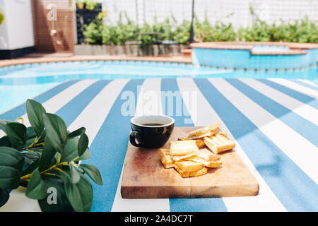 Tasse leckeren Kaffee und kleinen Toasts auf Holzbrett von Pool serviert Stockfoto