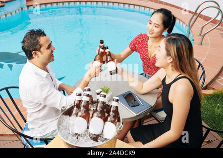 Gruppe der glücklichen asiatischen Menschen am Tisch sitzen, Swimmingpool und klirren Bier Flaschen Stockfoto