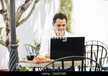 Positive junge Inder in Kaffee und Toast für breakdast bei Arbeiten am Laptop im Café im Freien Stockfoto