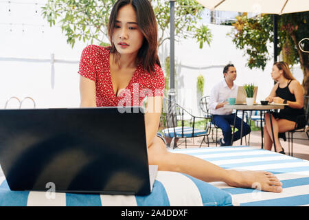 Ernsthafte barfuß Frau sitzt auf striped Chaise-lounge am Pool und Arbeiten am Laptop Stockfoto