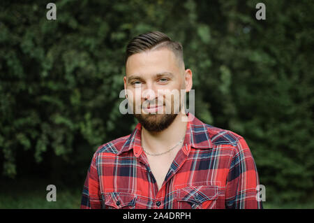 Nahaufnahme, Porträt junger bärtiger Mann im roten Hemd in die Kamera schaut mit einem Lächeln im Park auf Bäumen Hintergrund Stockfoto