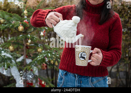 Frau giesst Tee mit einer Teekanne in Wasserglas. Weihnachtsbaum im Vordergrund. Teetasse mit Weihnachtsschmuck. Dampf aus Tasse Tee. Winter Stockfoto