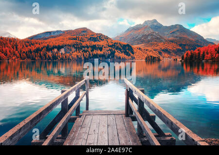 Hölzerne Seebrücke im Herbst den Silsersee (Silsersee) in den Schweizer Alpen. Snowy Mountains und Orangenbäumen im Hintergrund. Schweiz, Maloja Region Oberengadin. Landschaftsfotografie Stockfoto