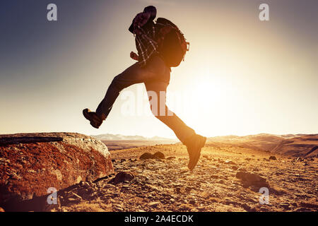 Wanderer Mann springt mit grossen Rucksack zu einem Rock gegen Berge und den Sonnenuntergang Sonne Stockfoto