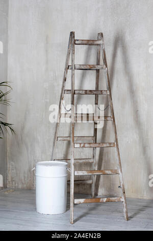 Eine hölzerne Stehleiter mit einem Barrel Farbe steht auf dem Stockwerk gegenüber einer strukturierten graue Wand. Stockfoto