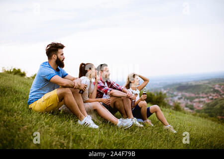 Gruppe der glückliche junge Freunde auf dem auf dem Gras an einem Hügel und trinken Apfelwein sitzen Stockfoto