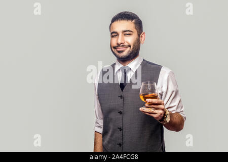 Portrait von gut aussehenden bärtigen Mann im weißen Hemd und Weste, die hielten Glas Whiskey und Kamera mit glücklichen Gesicht und Lächeln. Stockfoto
