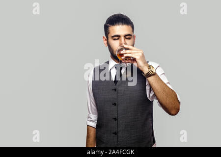 Portrait von gut aussehenden Bärtigen brünett Mann im weißen Hemd und Weste, die hielten Glas Whiskey und trinken mit geschlossenen Augen. indoor Studio Stockfoto