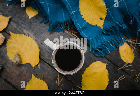 Auf einem alten Holztisch im Herbst Park ist eine Tasse Kaffee - Kaffee. Eine blaue warme karierte Schal ist mit gelben Blätter verstreut und Tannenzapfen. Ansicht von oben Stockfoto