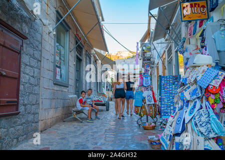 Hydra, griechische Insel - 20. Juli 2019; Drei junge Frauen zu Fuß entfernt, während die kleinen Jungen auf der Bank gegenüber souviners auf Anzeige in schmale Gasse sitzen. Stockfoto