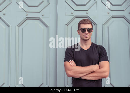 Athlet Mann im Sommer in der Stadt, voller Zuversicht in die Sonnenbrille steht, Hintergrund hölzerne Tür Tor, Schwarz T-Shirt. Eine ernsthafte überzeugter Kerl mit seinen Armen Stockfoto