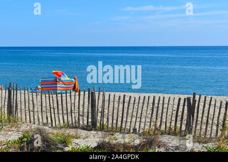 Bunten Sonnenschirm und gestreifte Windschutz hinter einem Zaun auf einen Kilometer langen Sandstrand im Süden von Frankreich. Ruhige blaue Meer trifft, dem hellen Himmel Stockfoto