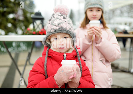 Zwei adorable Schwestern trinken heiße Schokolade auf den traditionellen Weihnachtsmarkt in Vilnius, Litauen. Kinder mit Süßigkeiten, Bonbons und Lebkuchen auf Stockfoto