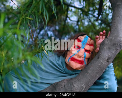 Eine verschleierte Person mit einem gruseligen Halloween Mask blickt zwischen den Ästen eines Baumes. Stockfoto