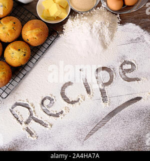 Das Wort Rezept geschrieben in Mehl auf einem dunklen Holztisch mit frisch gebackenen Muffins und Zutaten. Stockfoto