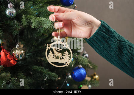 Hände im grünen Pullover halten und Holzspielzeug Hirsche auf künstlichen grünen Weihnachtsbaum extreme Ansicht schließen hängen Stockfoto