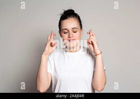 Eine aufgeregte junge Frau mit gekreuzten Fingern macht ein Wunsch, schloss die Augen und hofft, dass alles wahr werden. Auf einem weißen Hintergrund. Stockfoto