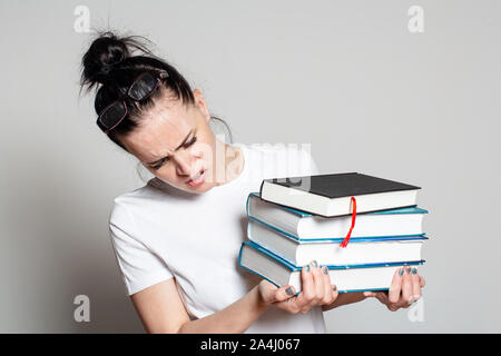 Schockiert, junge Studentin mit Brille auf dem Kopf hält einen Stapel von Büchern in den Händen, schaut sie verwundert an. Auf weissem Hintergrund. Stockfoto