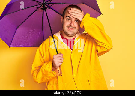 Junger Mann mit regenmantel Holding lila Schirm stehend über isolierte gelber Hintergrund mit Hand betonte am Kopf, schockiert mit Scham und surpris Stockfoto