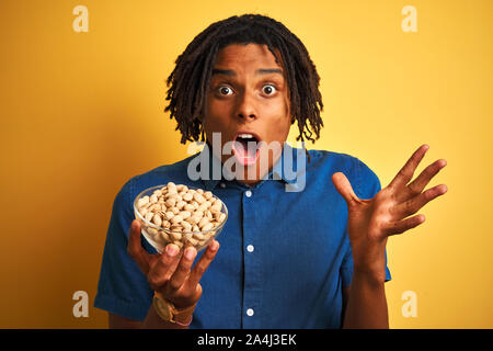 Junge afro-amerikanische Mann mit Dreadlocks essen Pistazien über isolierte gelben Hintergrund sehr glücklich und aufgeregt, Sieger Ausdruck victor Feiern Stockfoto