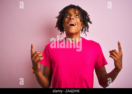 Afro-amerikanische Mann mit Dreadlocks tragende t-shirt stehend über isolierte rosa Hintergrund erstaunt und überrascht mit der Suche und zeigt mit Fingern und Stockfoto