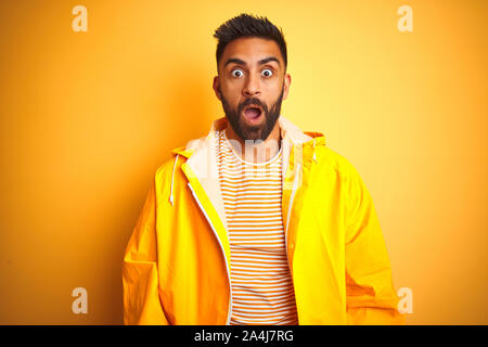 Junge indische Mann mit regenmantel stehend über isolierte gelben Hintergrund Angst und mit Erstaunen Ausdruck, Angst und aufgeregt Gesicht schockiert. Stockfoto