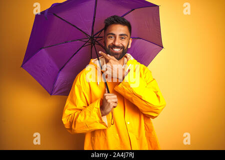 Junge indische Mann mit Regenmantel und lila Schirm über isolierte gelben Hintergrund sehr glücklich zeigte mit Hand und Finger Stockfoto