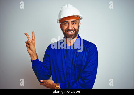 Hübscher indischer Arbeitnehmer Mann einheitliche und Helm tragen über isoliert weißer Hintergrund lächelnd mit glücklichen Gesicht in die Kamera winking tun Sieg unterzeichnen. Stockfoto