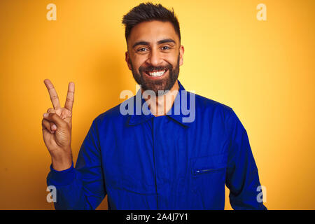Hübscher indischer Arbeitnehmer Mann in Uniform über isolierte gelben Hintergrund lächelnd mit glücklichen Gesicht in die Kamera winking tun Sieg unterzeichnen. Anzahl tw Stockfoto