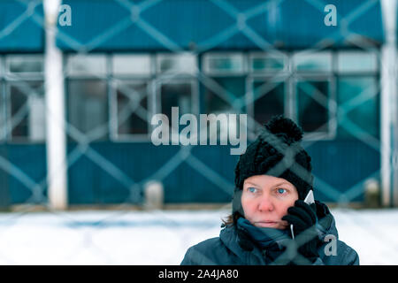 Gestörte Frau Gespräch am Handy auf der Straße hinter der Chain-link Fence, Hindernis auf kalten Wintertag symbolisiert, selektiver Fokus Stockfoto