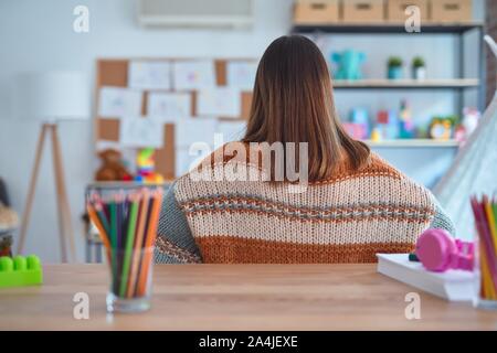 Junge schöne Lehrerin Frau mit Pullover und Gläsern sitzen am Schreibtisch im Kindergarten stehen nach hinten weg schauen mit den Armen am Körper Stockfoto