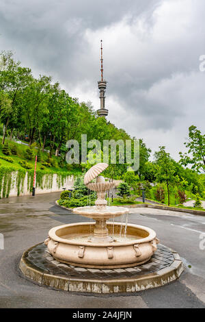 Almaty Kok Tobe Blue Hill Mountain Park, mit Blick auf einen Brunnen mit Fernsehturm im Hintergrund an einem nebligen und trüben Tag Stockfoto