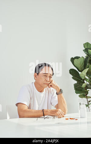 Ältere Menschen am Tisch sitzen, schlechte Kopfschmerzen, Grimassen, Einnahme von Medizin.? Stockfoto