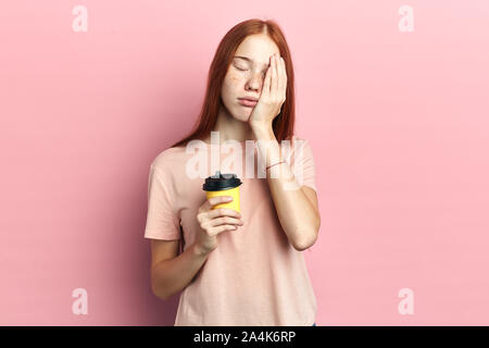 Müde, schläfrig Frau mit geschlossenen Augen deckt die Hälfte der Fläche mit Palm, hat traurige Ausdruck, hält Einweg Tasse trinken, die Koffein enthalten. isolierte Klemme Stockfoto