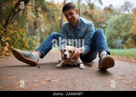 Fröhlicher junger stattlicher Mann sitzen auf der Straße mit seinem adorle Pet, genießen die Zeit an der frischen Luft. in voller Länge Foto. Freundschaft Stockfoto