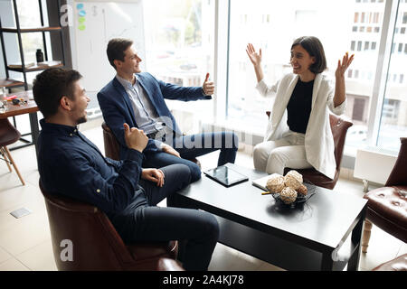 Zwei fröhliche stattlichen Geschäftsleute zeigen Daumen bis zu ihren weiblichen Kollegen, am Tisch mit ausgestreckten Armen sitzt, unseren herzlichen Glückwunsch! Stockfoto