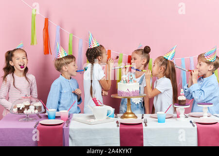 Lustige kleine Jungen und Mädchen blasen in die Partei Hörner, unterhaltsam, isolierte rosa Hintergrund, Urlaub, festliche Zeit Stockfoto