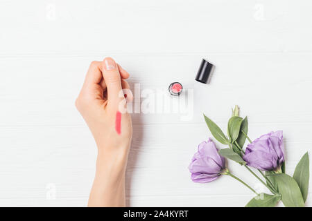 Frau testen rosa Lippenstift auf Ihre Hand neben lila Blumen auf weißem Holz- Tabelle, Ansicht von oben. Stockfoto