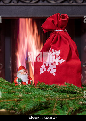 Ein wenig Santa Claus Spielzeug Abbildung steht auf grüne Zweige Fichte neben einer roten Geschenkbeutel. Im Hintergrund ist ein Feuer im Kamin. Gemütliche Einrichtung Stockfoto