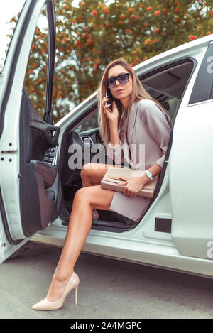 Frau steigt aus dem Auto sommer herbst Stadt. Taxi Business Class. In der Hand Handy, Carsharing, Mietwagen, rosa Anzug Schuhe mit hohen Absätzen. Gebräunten schlanken Mädchen Stockfoto