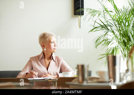 Reife Geschäftsfrau mit kurzen blonden Haare weg schauen, während am Tisch im Büro arbeiten Stockfoto