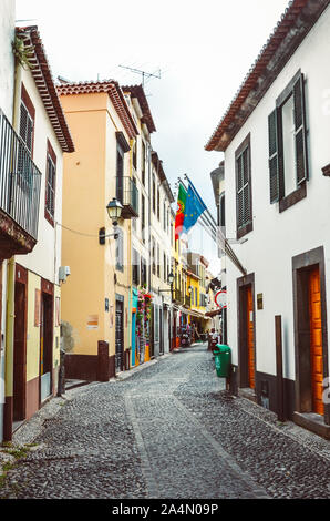 Funchal, Madeira, Portugal - 21.September 2019: eine gepflasterte Gasse in der Altstadt der Hauptstadt Madeiras bei bedecktem Himmel. Historische Gebäude mit winkenden Portugiesisch und EU-Flaggen. Stadtzentrum. Stockfoto