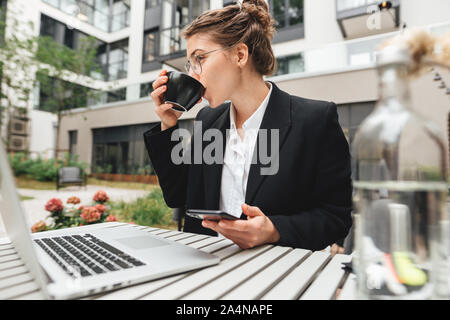 Young Business Frau mit Brille im Cafe mit Laptop und Kaffee zu trinken. Geschäftsfrau trinken Kaffee im Cafe und halten Smartphone Stockfoto