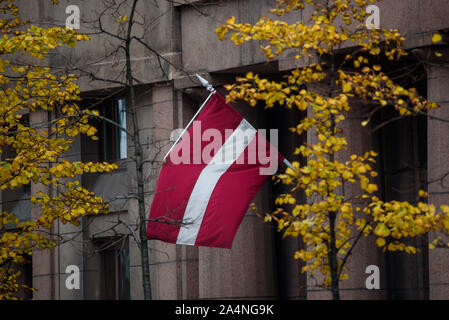 RIGA, Lettland. 15. Oktober, 2019. Flagge Lettland vor der Fassade zwischen zwei Bäumen mit gelben Blätter gehängt. Stockfoto