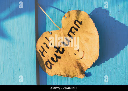 "Hallo Herbst!' - eine Inschrift auf einem gelben Blatt, das von einem Baum auf einem blauen Holz- Oberfläche fiel Stockfoto