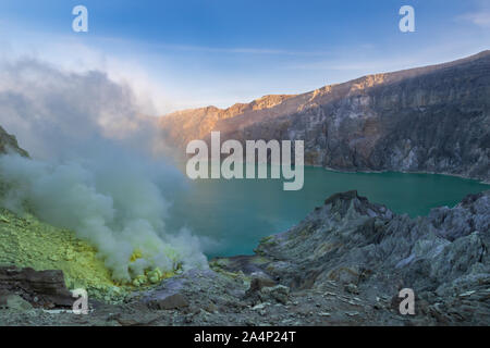 Säure auf dem Kratersee des Ijen, Ostjava, Indonesien. Rauch und Schwefel Ablagerungen am Ufer aus vulkanischen Schloten. Blue Sky hinter weit Kraterwand. Stockfoto